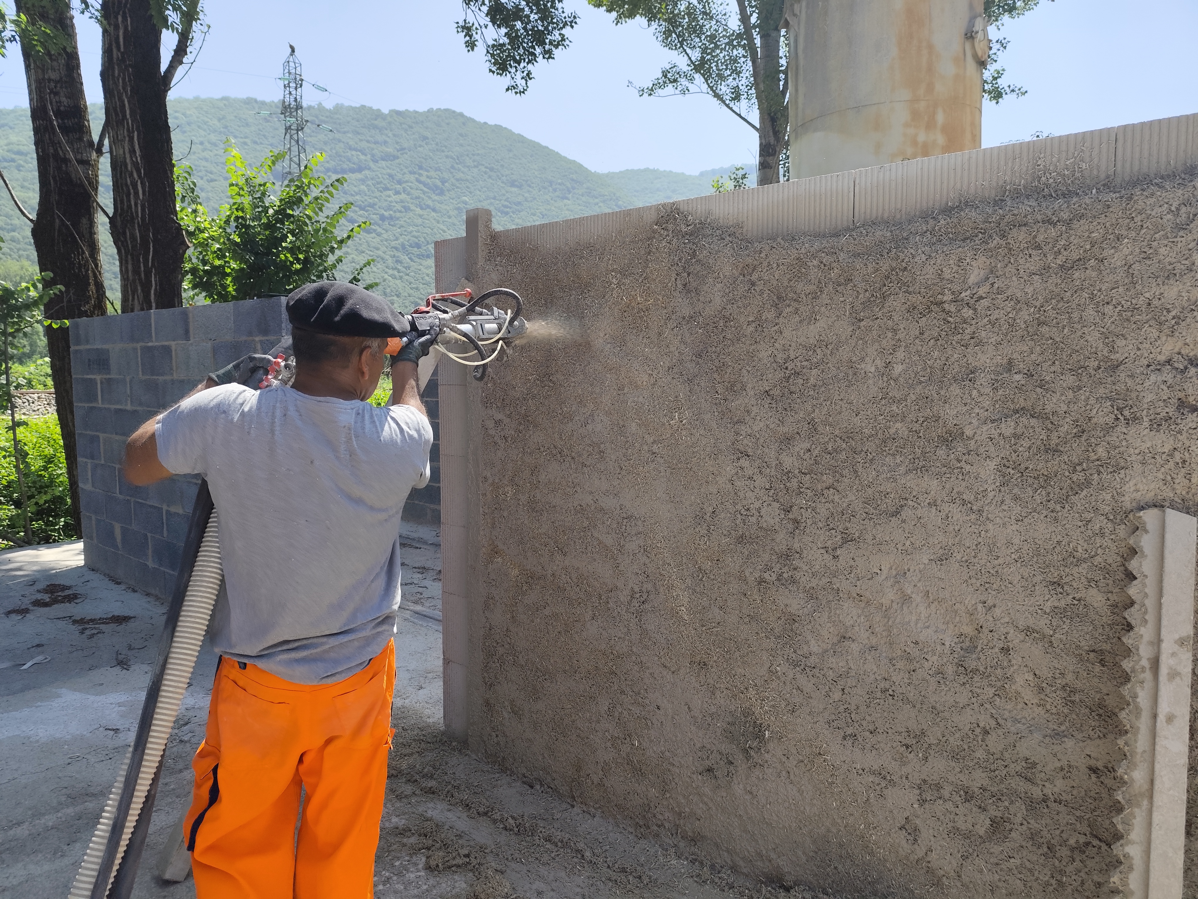 Un ouvrier en tenue orange pulvérise du mortier sur un mur en béton à l'aide d'une machine en extérieur, entouré d'arbres et de collines en arrière-plan.