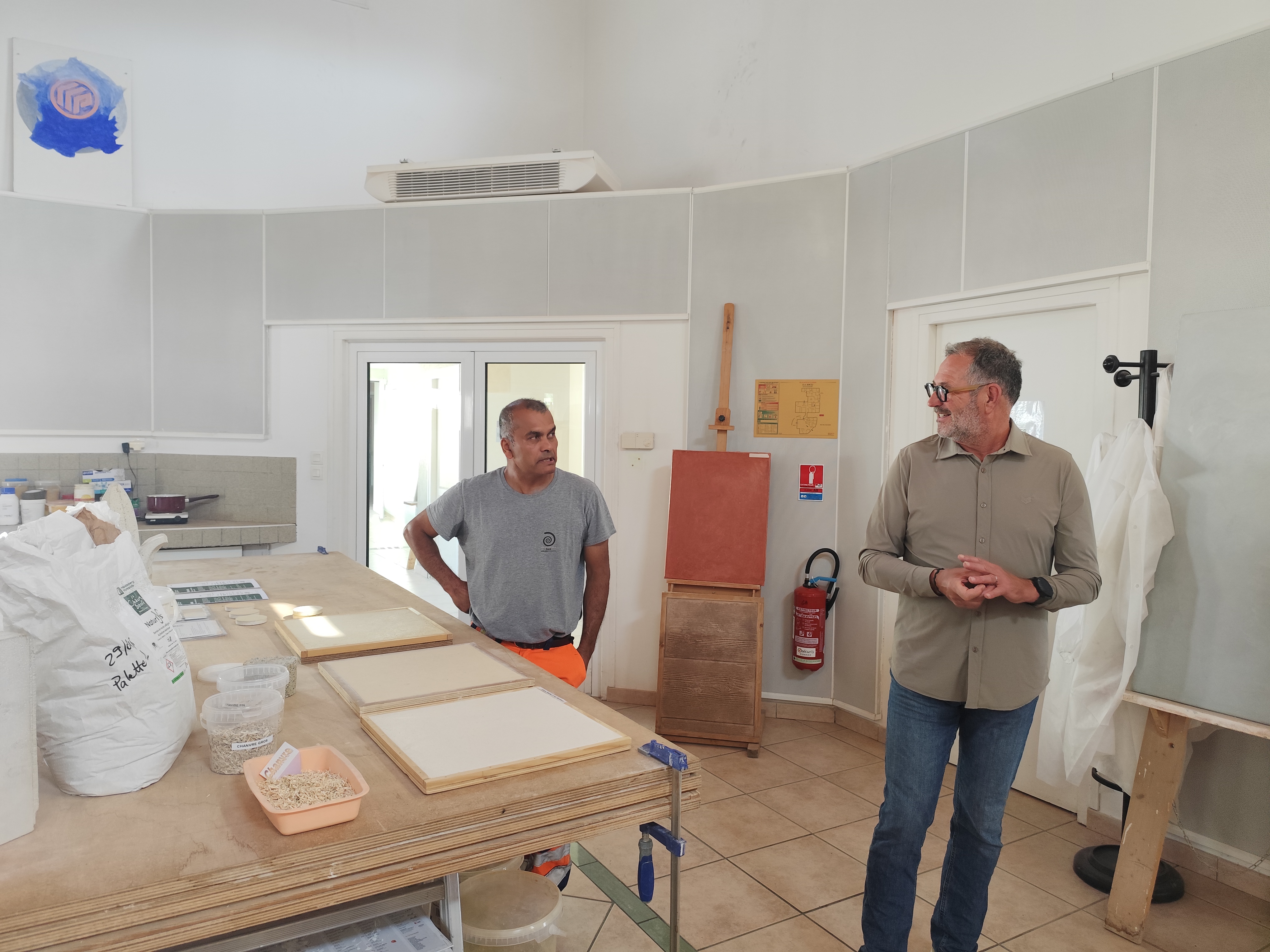 Deux hommes discutent dans un atelier lumineux. À gauche, un homme en t-shirt gris et pantalon orange se tient près de sacs de matières premières et cadres en bois sur une table. À droite, un homme en chemise beige parle, face à l'autre. En arrière-plan, murs blancs, une porte vitrée, et des outils accrochés dont une pelle et un extincteur.